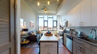 This 1-bedroom, 1-bathroom unit showcases a compelling blend of industrial and contemporary design throughout its single-floor layout. The open-concept living and kitchen area is anchored by a striking exposed concrete ceiling, warm hardwood-style flooring, and oversized black-framed windows that flood the space with natural light and offer urban views of the surrounding neighborhood.

The kitchen is well-appointed with stainless steel appliances, including a range, over-the-range microwave, and refrigerator. Two-tone cabinetry pairs white upper cabinets with wood-toned lower cabinets, complemented by a subtle tile backsplash and stone-look countertops. A freestanding island with a white surface provides additional prep space and casual seating for two.

The living area is generously sized, with tall ceilings that enhance the sense of volume and an abundance of natural light streaming through floor-to-ceiling windows. The bedroom continues the industrial aesthetic with an exposed concrete accent column, large windows with urban views, and ample room for furnishings. The bathroom features a wood-toned vanity with an undermount sink, stone-look countertop, and hardwood-style flooring, completing the cohesive modern design scheme throughout the unit.