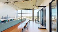 Located at 754 E 13th Ave in Eugene, Oregon, this thoughtfully designed coworking space occupies a single floor and offers a compelling blend of modern industrial architecture and nature-inspired aesthetics. Exposed concrete ceilings with integrated track lighting lend an urban, loft-like character to the space, while a herringbone-patterned light wood floor adds warmth underfoot.

Expansive floor-to-ceiling windows dominate one full wall, flooding the interior with natural light and offering elevated panoramic views of the Eugene cityscape and surrounding mountains — a standout feature of this space. Long communal worktables with teal acoustic privacy dividers provide ample open seating for focused, individual work or collaborative sessions. Seating options vary throughout, including bar-height stools and standard chairs to accommodate different working styles.

A nature-themed forest mural wraps across interior walls, creating a visually striking backdrop that echoes the Pacific Northwest setting. For more private work, glass-enclosed meeting pods with sliding doors offer a quiet retreat without sacrificing visual connectivity. A cozy lounge area with accent chairs adds a relaxed atmosphere near the windows. The overall ambiance is bright, modern, and professional — well-suited for productive work in an inspiring environment.