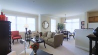 This well-appointed single-floor residence offers a thoughtfully designed open-concept layout that seamlessly connects the living and dining areas into one cohesive, light-filled space. Large windows line the walls, flooding the interior with abundant natural light and creating an airy, welcoming atmosphere throughout.

The living area is anchored by a plush, neutral-toned sofa complemented by a sleek dark coffee table and coordinating side tables, all resting atop soft wall-to-wall carpeting. A decorative sunburst mirror serves as a focal point on the wall, adding a refined artistic touch to the space. Crown molding runs along the ceiling line, lending a polished, classic architectural detail that elevates the overall aesthetic.

Transitioning naturally into the dining area, a round dark-wood dining table is surrounded by elegant upholstered chairs beneath a pendant light fixture, creating an intimate setting ideal for entertaining. Lush indoor greenery adds warmth and a touch of nature to the corner of the space.

The neutral color palette—featuring soft beiges, creams, and warm whites—creates a timeless and sophisticated ambiance. Carefully placed floral arrangements in rich, warm tones provide vibrant accents throughout. The overall design strikes a balance between classic elegance and comfortable, livable style.