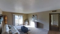 This single-floor space centers around a generously sized living room that offers a comfortable and functional layout. The room is finished with wall-to-wall carpeting in a neutral tone, contributing to a warm and inviting atmosphere. A textured popcorn ceiling adds a classic architectural detail overhead, while the walls are painted in a soft, earthy taupe that complements the overall color palette.

Natural light floods the room through a large sliding glass door positioned along one wall, offering a view of an outdoor area and brightening the interior throughout the day. The sliding door also provides convenient outdoor access, enhancing the sense of openness within the space.

A low-profile credenza or media console sits against one wall beneath a bold geometric artwork, serving as a natural focal point. The living area accommodates a patterned accent chair and a zebra-print area rug layered beneath a tufted ottoman, adding visual interest and texture to the space. White painted trim frames the walls and doorways, providing clean contrast against the warmer wall tones. Overall, the space presents a relaxed yet stylish environment suited for everyday living.