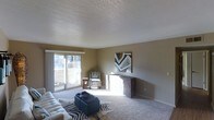 This single-floor space centers around a generously sized living room that offers a comfortable and functional layout. The room is finished with wall-to-wall carpeting in a neutral tone, contributing to a warm and inviting atmosphere. A textured popcorn ceiling adds a classic architectural detail overhead, while the walls are painted in a soft, earthy taupe that complements the overall color palette.

Natural light floods the room through a large sliding glass door positioned along one wall, offering a view of an outdoor area and brightening the interior throughout the day. The sliding door also provides convenient outdoor access, enhancing the sense of openness within the space.

A low-profile credenza or media console sits against one wall beneath a bold geometric artwork, serving as a natural focal point. The living area accommodates a patterned accent chair and a zebra-print area rug layered beneath a tufted ottoman, adding visual interest and texture to the space. White painted trim frames the walls and doorways, providing clean contrast against the warmer wall tones. Overall, the space presents a relaxed yet stylish environment suited for everyday living.