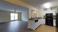 This single-floor home offers a comfortable and practical layout designed for everyday living. The kitchen is outfitted with white cabinetry, dark countertops, a built-in dishwasher, and a sink positioned along a peninsula that connects seamlessly to the rest of the living space. The open-concept design allows for easy flow between the kitchen and the adjoining living area, creating a cohesive and sociable environment. Warm hardwood-style flooring extends throughout the main areas, adding a consistent and inviting aesthetic underfoot. The living room is generously sized, filled with natural light streaming through a large window, and features a ceiling fan that adds both function and a classic touch to the space. Neutral, light-toned walls throughout keep the atmosphere bright and versatile, easily adaptable to a variety of interior styles. A doorway visible from the kitchen hints at additional rooms beyond, suggesting a well-organized floor plan. The textured ceilings and clean architectural lines throughout contribute to a straightforward, move-in-ready character. Overall, this single-story home balances practicality with comfort, making it a welcoming and functional living environment.