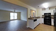 This single-floor home offers a comfortable and practical layout designed for everyday living. The kitchen is outfitted with white cabinetry, dark countertops, a built-in dishwasher, and a sink positioned along a peninsula that connects seamlessly to the rest of the living space. The open-concept design allows for easy flow between the kitchen and the adjoining living area, creating a cohesive and sociable environment. Warm hardwood-style flooring extends throughout the main areas, adding a consistent and inviting aesthetic underfoot. The living room is generously sized, filled with natural light streaming through a large window, and features a ceiling fan that adds both function and a classic touch to the space. Neutral, light-toned walls throughout keep the atmosphere bright and versatile, easily adaptable to a variety of interior styles. A doorway visible from the kitchen hints at additional rooms beyond, suggesting a well-organized floor plan. The textured ceilings and clean architectural lines throughout contribute to a straightforward, move-in-ready character. Overall, this single-story home balances practicality with comfort, making it a welcoming and functional living environment.
