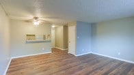 This single-floor space showcases an open-concept design that seamlessly connects the main living area to the kitchen, creating a sense of openness and easy flow throughout. Rich hardwood flooring runs across the living space, adding warmth and a timeless aesthetic to the interior. The textured ceiling adds a classic finishing touch, while a ceiling fan with a light fixture provides both comfort and illumination to the main living area.

The kitchen features a raised pass-through counter that overlooks the living space, making it ideal for casual dining or entertaining. Appliances, including a refrigerator, are visible within the kitchen area, complementing the functional layout. Soft, neutral-toned walls in muted blues and greens lend the space a calm and inviting atmosphere.

Interior doorways lead to additional rooms or utility spaces, suggesting a well-organized floor plan. Clean white baseboards and trim throughout provide a crisp contrast to the walls and flooring, adding a polished, well-maintained look. Overall, the space offers a practical and comfortable single-level layout that balances simplicity with functionality.