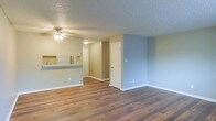 This single-floor space showcases an open-concept design that seamlessly connects the main living area to the kitchen, creating a sense of openness and easy flow throughout. Rich hardwood flooring runs across the living space, adding warmth and a timeless aesthetic to the interior. The textured ceiling adds a classic finishing touch, while a ceiling fan with a light fixture provides both comfort and illumination to the main living area.

The kitchen features a raised pass-through counter that overlooks the living space, making it ideal for casual dining or entertaining. Appliances, including a refrigerator, are visible within the kitchen area, complementing the functional layout. Soft, neutral-toned walls in muted blues and greens lend the space a calm and inviting atmosphere.

Interior doorways lead to additional rooms or utility spaces, suggesting a well-organized floor plan. Clean white baseboards and trim throughout provide a crisp contrast to the walls and flooring, adding a polished, well-maintained look. Overall, the space offers a practical and comfortable single-level layout that balances simplicity with functionality.