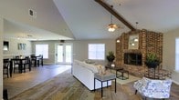 This single-story space makes a strong architectural impression with its dramatic vaulted ceilings, accented by an exposed wooden beam that runs diagonally across the roofline. A ceiling fan with a light fixture hangs from the peak, complementing a pair of pendant lights that add warmth to the open layout. The living area is anchored by a full-height brick fireplace, which serves as a striking focal point and adds a rustic, textured contrast to the otherwise neutral, light-toned walls. The flooring transitions between areas, featuring a patterned area rug beneath the seating arrangement and hard-surface flooring extending toward the entry zone. Natural light flows generously through multiple windows fitted with horizontal blinds, as well as a set of French doors that open to an exterior view, enhancing the sense of openness. The seating area is furnished with a white sofa and modern accent tables, creating an inviting and functional gathering space. A secondary area near the French doors offers additional versatility. The overall ambiance balances casual comfort with distinctive architectural character.