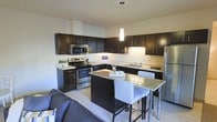 This single-floor residence showcases a well-designed open-concept layout that seamlessly integrates the kitchen and living areas. The kitchen is outfitted with rich, dark espresso cabinetry that extends to the upper walls, providing generous storage throughout. Stainless steel appliances, including a refrigerator, electric range, and over-the-range microwave, lend a sleek, contemporary aesthetic. Light granite countertops contrast elegantly with the dark cabinetry, and the space is anchored by a central island that doubles as a breakfast bar, accommodating two bar-height chairs — ideal for casual dining or entertaining. A single pendant light hangs above the island, providing focused task lighting while contributing to the modern design scheme. Under-cabinet lighting further enhances the kitchen's functionality. Light-toned flooring runs throughout the space, complementing the overall neutral color palette and contributing to a bright, airy atmosphere. Adjacent to the kitchen, the living area is partially visible, hinting at a cohesive and comfortable layout. The open flow between spaces makes this residence feel spacious and practical for everyday living.