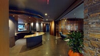 This thoughtfully designed single-floor amenity space showcases a striking blend of warm wood accents, polished concrete floors, and contemporary furnishings. At the heart of the space is a curved reception desk topped with a richly veined stone countertop, complemented by an eye-catching slatted wood ceiling detail overhead that adds architectural depth and warmth.

A standout feature is the curved wine storage room, enclosed by dramatic floor-to-ceiling vertical wood slat partitions that offer both visual intrigue and a sense of intimacy. Inside, illuminated wine display cabinetry lines the walls alongside a dining table, creating an ideal setting for private gatherings.

The full kitchen is outfitted with professional-grade stainless steel appliances, including a built-in double oven, a large French door refrigerator, and a gas cooktop set into a dark granite countertop — all framed by slatted wood ceiling panels that carry the design language throughout.

A generous lounge area features curved built-in seating, clustered dining tables, and large sliding glass doors that open to an outdoor pool area, inviting natural light and seamless indoor-outdoor connectivity. Lush tropical potted plants are positioned throughout, adding a vibrant, organic contrast to the dark, moody palette. The overall ambiance is upscale, cohesive, and welcoming.