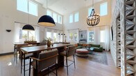 This well-appointed clubroom is designed to impress, showcasing dramatic double-height ceilings that create an expansive, airy atmosphere throughout the open-plan layout. Wide-plank hardwood flooring flows seamlessly across the entire space, anchoring the various functional zones with warmth and cohesion.

The central lounge area is anchored by a generous sectional sofa in deep olive green, accented with rust-toned throw pillows and layered over a vintage-style area rug. A textured geometric accent wall serves as the backdrop for a large-format flat-screen television, while a statement beaded chandelier overhead adds an artistic focal point. Large windows dressed in sheer white curtains flood the room with natural light and offer views of the surrounding outdoor environment.

Adjacent to the lounge, a solid wood high-top dining table with upholstered metal-frame chairs provides an ideal gathering spot beneath a bold oversized dome pendant light. A built-in kitchenette nearby features sleek navy cabinetry, rich walnut wood accents, a dramatic dark tile backsplash, and stainless steel appliances—both functional and visually striking.

A pool table occupies one corner of the space, while a cozy game nook lined with sculptural three-dimensional wall panels offers intimate seating around a bistro-style game table. The overall ambiance is modern, curated, and inviting—ideal for socializing and relaxation.