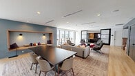 This expansive, contemporary amenity space offers a thoughtfully designed, multi-level layout connected by sleek glass-railed staircases and open mezzanine walkways. Light hardwood flooring flows throughout, complementing the clean white ceilings fitted with recessed lighting and linear HVAC diffusers that maintain a polished, modern aesthetic.

The main lounge area is generously proportioned, furnished with an eclectic mix of gray upholstered armchairs, plush sofas, and bold accent pieces in deep red and charcoal tones, all anchored by large area rugs. Floor-to-ceiling windows wrap around key areas of the space, flooding the interiors with abundant natural light and offering unobstructed views of the surrounding urban landscape.

A dedicated media lounge features a large wall-mounted flat-screen television framed by warm wood built-in shelving and cabinetry, with comfortable sectional seating and tufted ottomans arranged for an immersive viewing experience. Elsewhere, intimate seating nooks with high-backed chairs provide quieter retreats for focused work or conversation.

The mezzanine level offers additional seating along the perimeter windows, with bar-height tables and stools taking advantage of the elevated views. A dramatic pendant light fixture cascades through the double-height atrium, serving as a striking focal point that ties the levels together. The overall ambiance is sophisticated, open, and inviting.