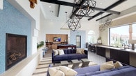 The Game Room at 2510 West Ranch in Carlsbad is a thoughtfully designed communal space that seamlessly blends comfort and entertainment. The room commands immediate attention with its soaring vaulted ceilings accented by dark exposed beams, from which striking geometric black pendant lights are suspended, creating a dramatic and modern aesthetic throughout.

The layout is open and inviting, anchored by plush navy blue sofas arranged over a bold striped area rug, providing a comfortable lounge setting for socializing. At the heart of the space sits a pool table, positioned beneath a wall-mounted flat-screen television and a warm wood-paneled accent wall, defining a dedicated recreation zone. The flooring transitions between hardwood-style planks and decorative patterned tile, adding visual interest and delineating activity areas.

Natural light pours in through arched windows and glass doors, illuminating the crisp white walls and vibrant blue patterned tile feature wall near the entrance. Along the perimeter, a sleek counter-height bar and seating area offers additional space for gathering. Lush greenery is thoughtfully placed throughout, lending a fresh, welcoming ambiance. The overall atmosphere is energetic yet refined, making this game room an ideal space for recreation and community connection.