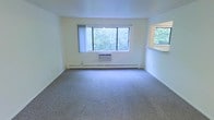 This single-floor space offers a clean and functional layout designed for comfortable everyday living. The main living area is finished with wall-to-wall carpet in a neutral gray tone, providing a soft and practical underfoot surface. Large windows dominate the exterior wall, inviting generous amounts of natural light and framing pleasant views of surrounding greenery. A wall-mounted air conditioning unit sits beneath the windows, ensuring climate control during warmer months, while baseboard heating runs along the perimeter of the room for year-round comfort. The walls are finished in a soft, light yellow-white tone, contributing to a bright and airy atmosphere. A secondary window on the adjacent wall adds additional natural light and ventilation to the space. The overall ambiance is simple, clean, and well-lit, making the space versatile and easy to personalize. The open layout of the living area allows for flexible furniture arrangements to suit a variety of needs and preferences.