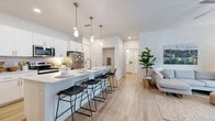 This well-appointed 2-bedroom, 2-bathroom residence in Phoenix, AZ offers a thoughtfully designed single-level layout that maximizes functionality and style. Light wood-look flooring flows throughout the open-concept living and kitchen areas, creating a cohesive, airy feel. The kitchen is a standout feature, showcasing white shaker cabinetry, stainless steel appliances, a large peninsula with bar seating, and sleek pendant lighting overhead — ideal for both cooking and entertaining. The living room is bright and welcoming, with a sliding glass door that opens to a private balcony offering views of lush landscaping and palm trees. Both bedrooms are carpeted for comfort and include dedicated closets with built-in shelving. The primary bedroom features a ceiling fan and generous walk-in closet. The primary bathroom is finished with a dual-sink white vanity, light wood-tone flooring, and a glass-enclosed walk-in shower. A dedicated in-unit laundry area houses a full-size washer and dryer. Two additional closets provide ample storage throughout the home. The overall design is clean and contemporary, with neutral tones and recessed lighting that contribute to a bright, modern ambiance throughout.