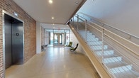 The lobby at Roebling Lofts showcases a thoughtful fusion of industrial heritage and contemporary design. Polished concrete floors extend across the open, airy entry space, reflecting light and contributing to the clean, minimalist aesthetic. Exposed brick walls on one side preserve the building's raw, loft-style character, while crisp white walls and recessed ceiling lighting create a bright, welcoming atmosphere.

A striking open staircase with a sleek metal frame and cable railing system commands attention on one side of the lobby, offering both visual interest and a sense of vertical openness. Floor-to-ceiling glass entry doors allow abundant natural light to flood the space, enhancing its expansive feel and providing clear sightlines to the street beyond.

A pair of modern lounge chairs adds a subtle residential touch, offering a comfortable seating area within the entrance. The overall design language is clean and purposeful — industrial materials are balanced by refined finishes, creating a lobby that feels both functional and architecturally distinctive. The space effectively sets the tone for the loft-style residences found throughout Roebling Lofts.