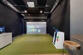 Located at The Sylvan at Painted Tree in McKinney, Texas, this dedicated indoor golf simulator room delivers a premium recreational experience within a compact, well-appointed space. The room is divided into two distinct functional zones that transition seamlessly from one to the other.

The simulation area dominates one side of the room, featuring a full synthetic turf hitting surface fitted with tee stations and a large high-definition projection screen powered by an aboveGOLF system. A ceiling-mounted projector delivers immersive virtual course visuals, while dark-painted walls and black curtained enclosures minimize glare and maintain focus on the screen. Club storage and ball racks are conveniently positioned at the edge of the turf for easy access.

The lounge area transitions to warm wood-look flooring, offering a welcoming space to relax and socialize. A tufted caramel-toned sofa, industrial-style pub table with live-edge stools, and a service counter equipped with a mini-refrigerator and monitor round out the amenities. Glass entry doors allow natural light into the lounge zone, providing an open, airy contrast to the moody simulator bay. Recessed lighting and track fixtures illuminate the entire space effectively. This room offers a polished, all-in-one golf and social experience for residents of the community.