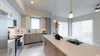 This well-appointed single-floor residence showcases a clean, contemporary design with a bright and airy ambiance throughout. The open-concept living and kitchen area serves as the heart of the space, where large sliding glass doors and floor-to-ceiling vertical blinds flood the room with natural light while offering views of the outdoors. Light-toned large-format tile flooring spans the main living areas, contributing to the seamless, modern aesthetic.

The kitchen is thoughtfully designed with two-tone cabinetry in white and warm wood-grain finishes, a white subway tile backsplash, and stainless steel appliances including a dishwasher. A generous center island with a crisp quartz countertop provides ample workspace and casual seating, accented by elegant pendant lighting with brushed brass fixtures. A dedicated pantry or storage cabinet adds practical functionality to the kitchen layout.

The hallway features louvered bi-fold closet doors, light hardwood-style flooring, and flush ceiling lighting, maintaining the home's consistent, polished palette. A wall-mounted thermostat is conveniently located in this corridor. Throughout the space, white trim, panel-style doors, and soft neutral wall tones create a cohesive and inviting atmosphere ideal for modern living.