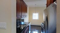 This single-floor space presents a thoughtfully designed kitchen and dining area that balances functionality with classic elegance. The kitchen is outfitted with floor-to-ceiling rich, dark wood cabinetry featuring raised-panel detailing and crown molding accents, offering generous storage throughout. Dark granite countertops provide a striking contrast against the warm cabinetry tones, while a stainless steel undermount sink with a modern arc faucet adds a refined touch. Stainless steel appliances, including a refrigerator and gas range, complete the kitchen's contemporary-classic aesthetic. The flooring throughout is polished dark tile, lending a sleek, high-end feel underfoot. The adjacent dining area is finished in soft, warm neutral tones and is anchored by a chandelier suspended from the ceiling, which adds an elegant focal point to the space. A window dressed with white blinds allows natural light to filter into the dining area, brightening the overall atmosphere. Crown molding details along the ceiling add architectural character throughout, contributing to a cohesive and polished interior.