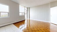 Located at 99 Tyndall Ave in Toronto's vibrant Parkdale neighbourhood, this compact studio unit offers a well-considered layout that maximizes every inch of its floor area. The open-concept kitchen and living room form the heart of the space, seamlessly connected to allow for flexible use. The kitchen is outfitted with stainless steel appliances — including a full-size refrigerator, stove, and dishwasher — complemented by white cabinetry, granite-style countertops, a classic subway tile backsplash, and marble-look tile flooring. Natural light pours into the kitchen through a large window overlooking the city skyline. The living area features warm, glossy parquet wood flooring that adds character and continuity throughout the space. Expansive floor-to-ceiling windows bathe the living room in abundant natural light while offering sweeping city views, and a sliding door provides direct access to the private balcony. The entryway includes a dedicated closet for storage. The bathroom is clean and functional, with a bathtub and tiled finishes. Baseboard heating runs throughout, ensuring year-round comfort. The overall aesthetic is clean and bright, with neutral grey walls and white trim creating a versatile backdrop.