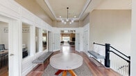 Located at 100 Cobblestone Dr in Coatesville, PA, this elegantly appointed single-floor community clubhouse offers a thoughtfully designed layout encompassing a living room, kitchen, and two hallways. The living room is the undisputed centerpiece, boasting dramatic double-height coffered ceilings, a striking floor-to-ceiling stacked stone fireplace, and an abundance of natural light streaming through oversized windows dressed with warm bamboo shades. A sophisticated globe chandelier and an arrangement of modern upholstered seating complete the inviting atmosphere. The bright, welcoming hallways feature white wainscoting, hardwood flooring, a built-in window seat, and glass-paneled office spaces that offer both transparency and functionality. The well-equipped kitchen includes rich wood cabinetry, dark granite countertops, and stainless steel appliances. Throughout the space, warm taupe walls, polished hardwood floors, and curated artwork create a cohesive and upscale aesthetic. A balcony overlooking a resort-style outdoor pool surrounded by mature trees adds a desirable outdoor dimension, rounding out this versatile and visually impressive community space.