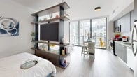 This thoughtfully designed studio occupies a single floor and makes efficient use of its open-plan layout. The main living area is anchored by a striking freestanding shelving unit that doubles as a room divider, seamlessly separating the sleeping zone from the living and dining areas while housing a large flat-screen TV. Light wood-style flooring runs throughout, complementing the bright, white walls and crisp modern aesthetic.

Floor-to-ceiling glass windows and a sliding door flood the interior with abundant natural light and open onto a balcony with expansive urban skyline views. The dining area is positioned to take full advantage of these views, with a compact bar-height table and seating situated near the glass perimeter. Adjacent to the living space, the kitchen is equipped with stainless steel appliances, contemporary cabinetry, and ample counter space.

The sleeping area benefits from a dedicated walk-in closet with built-in shelving and hanging storage. The bathroom features large-format tile flooring, a single vanity with under-cabinet storage, and a generously sized glass-enclosed walk-in shower with an overhead rain showerhead. Decorative artwork adds a finishing touch throughout the space, contributing to a vibrant yet cohesive interior character.