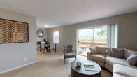 This single-floor residence is thoughtfully laid out to maximize comfort and functionality throughout. The living room serves as the heart of the home, finished with plush carpeting and neutral warm-gray walls that create a calm, cohesive atmosphere. A large sliding glass door floods the space with natural light and opens directly onto a private outdoor patio, offering pleasant greenery views beyond a wood-railed fence. The living area flows naturally into a separate dining room, which features its own window with blinds, soft carpeting, and ample space for a full dining set complemented by a side console.

The kitchen is efficiently designed with white cabinetry, light countertops, and tile flooring. It includes a double sink, electric range, and under-cabinet range hood, with a pantry closet providing additional storage.

The primary bedroom is generously sized, carpeted, and well-lit by a window framing outdoor greenery. It accommodates a king-sized bed comfortably alongside nightstands. A second bedroom offers similar neutral tones, carpet flooring, a closet, and a window with natural light, providing a versatile and inviting space.

The bathroom features tile flooring, a tub-shower combination, a white vanity with storage, and clean, neutral finishes throughout.