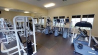 This fitness center is a dedicated single-floor exercise facility designed to support a comprehensive workout experience. The space is outfitted with a solid selection of equipment, including multiple elliptical machines and cardio steppers for aerobic training, as well as a cable weight machine for targeted strength exercises. Weight plates are neatly stacked and accessible, catering to users of varying fitness levels. A large wall mirror spans one side of the room, visually expanding the space and allowing users to monitor their form during workouts. The flooring consists of durable rubber matting, providing both safety and comfort underfoot. Natural light streams in through a row of windows along the exterior wall, creating a bright and energizing atmosphere. The walls are finished in a warm neutral tone, contributing to an inviting and focused environment. Overhead fluorescent lighting ensures the space remains well-lit at all times. The layout is thoughtfully arranged to allow ample room for movement between equipment, making the facility practical and easy to navigate for users of all experience levels.