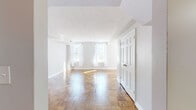 This single-floor residence offers a thoughtfully designed layout that balances classic charm with modern updates. Throughout the living and bedroom areas, warm parquet hardwood flooring laid in a distinctive basket-weave pattern adds timeless character and visual interest to every room. Crisp white walls and generous natural light streaming through multiple double-hung windows create a bright, airy ambiance across the space.

The spacious living area is open and welcoming, framed by elegant six-panel doors and defined architectural detailing including structural beams and clean crown molding. A secondary room continues the parquet flooring and features a built-in closet nook, offering practical storage within a well-proportioned space.

The kitchen has been updated with sleek white shaker cabinetry, granite countertops with bold natural veining, and a full suite of stainless steel appliances including a range, built-in microwave, and dishwasher. Light-toned tile flooring completes the clean, functional aesthetic.

The bathroom is finished with classic white subway tile wainscoting, mosaic tile flooring, and a pedestal-style vanity. A soaking tub, wall-mounted toilet, and a window providing natural ventilation round out this well-appointed room. Overall, the space presents a cohesive blend of traditional architectural details and contemporary finishes.