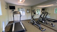 This fitness center is a dedicated space designed to support a well-rounded cardio workout routine. The room is furnished with key exercise equipment, including a treadmill and an elliptical trainer, positioned to make efficient use of the available floor space. A large floor-to-ceiling mirror lines one wall, visually expanding the room and allowing users to monitor their form during exercise. Carpeted flooring adds a layer of comfort underfoot, while framed artwork on the neutral-toned walls contributes a clean, welcoming aesthetic. Multiple windows allow natural light to filter into the space, offering pleasant views of the surrounding greenery and creating a bright, open atmosphere. A wall-mounted flat-screen television is positioned for easy viewing during workouts, adding a layer of entertainment to training sessions. Overhead lighting ensures the room remains well-lit at all times. The overall design of the fitness center is straightforward and functional, prioritizing ease of use and comfort. The combination of natural light, reflective surfaces, and thoughtfully arranged equipment makes this a practical and inviting space for daily exercise.