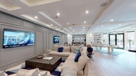 This single-level clubroom in College Station, Texas, is a thoughtfully designed social space that blends comfort and sophistication throughout. The main lounge area serves as the centerpiece, featuring an expansive sectional sofa in neutral tones accented with blue and teal throw pillows, arranged around a dark wood coffee table atop a geometric-patterned area rug. Multiple large flat-screen televisions are mounted along a striking gray wainscoted accent wall, creating an ideal setting for group gatherings or entertainment. Tray ceilings with recessed lighting add architectural depth and a refined ambiance to the space.

Beyond the lounge, a billiards table anchors a dedicated game area, offering additional recreational options for guests. The kitchen and bar area features a substantial marble-topped island, globe-style metallic pendant lights, a mosaic tile backsplash, and built-in appliances, providing a functional yet elegant space for hosting. A separate workspace nook includes a sleek marble-surfaced table, upholstered chairs, and matching pendant fixtures, ideal for focused use. Light wood-toned tile flooring flows consistently throughout, unifying the various zones while enhancing the overall bright, contemporary atmosphere of this well-appointed clubroom.