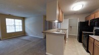 This single-story space showcases an open-concept layout that seamlessly connects the kitchen and living area. The kitchen is anchored by a peninsula counter with granite-style countertops, providing both additional prep space and a natural divider between the cooking and living zones. Oak-toned upper cabinetry offers ample storage, while an under-cabinet range hood and a stainless steel sink add functional detail. Tile flooring runs through the kitchen area, offering a practical and easy-to-maintain surface.

The adjacent living area features wall-to-wall carpeting, creating a warm and comfortable atmosphere. Neutral-toned walls throughout provide a versatile backdrop, complemented by a textured ceiling finish. A window in the living area allows for natural light to filter in, brightening the space.

Visible beyond the kitchen, a dedicated laundry area with built-in shelving offers added convenience. A hallway connects to additional rooms within the unit. The overall design is practical and functional, with a cohesive neutral color palette that lends a clean and straightforward aesthetic throughout.