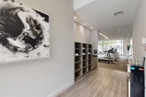 Located at 170 Century Square Drive in College Station, TX, this fitness center offers a spacious, single-floor exercise facility designed with both functionality and aesthetics in mind. The space is thoughtfully organized into distinct workout zones, beginning with a welcoming entryway finished in light wood-look flooring and accented by contemporary abstract wall art. Built-in shelving units provide convenient towel and accessory storage near the entrance, alongside a water station for easy access.

The main exercise floor transitions to a patterned carpet with blue accents, anchoring a comprehensive arrangement of equipment. A dedicated cardio zone features a row of treadmills and elliptical machines positioned along expansive floor-to-ceiling windows, flooding the space with natural light and offering open views of the surrounding streetscape. A separate cycling area accommodates multiple stationary and recumbent bikes, also benefiting from the bright, window-lined perimeter.

The strength training section is equally well-appointed, housing a multi-station cable machine, adjustable benches, a full dumbbell rack, barbells, and a variety of free weights and plates. Large wall mirrors amplify the sense of space and support proper form during workouts. Linear LED lighting runs across the ceiling throughout, providing consistent, even illumination. Mounted flat-screen televisions are positioned throughout the facility for added convenience during workouts.