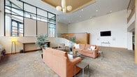 Located at 1201 E Market St in Akron, Ohio, this well-appointed lobby lounge is a welcoming communal space defined by its striking architectural character. Soaring double-height ceilings create an airy, expansive atmosphere, while a dramatic wall of floor-to-ceiling windows floods the interior with abundant natural light and provides sweeping views of the surrounding streetscape and neighboring structures.

The lounge area is thoughtfully arranged with warm terracotta-toned sofas and accent chairs positioned around low-profile coffee tables, fostering a relaxed yet polished environment. Textured neutral carpeting grounds the space, complementing the warm wood accents found throughout, including a curved wood-paneled feature wall that encloses a gracefully sweeping staircase with glass and stainless steel railings — a true architectural centerpiece. Potted indoor plants add organic touches of greenery.

A cluster of pendant globe light fixtures is suspended from a recessed ceiling tray, providing warm ambient illumination alongside recessed downlights. A wall-mounted flat-screen display and a sleek console table offer additional functionality. The space also connects to a corridor leading to further amenities. The overall ambiance is professional, modern, and inviting — ideal as a gathering or reception area.