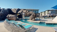 This three-floor residential community is defined by its exceptional outdoor amenity space, centered around a striking resort-style pool complex that serves as the heart of the development. The centerpiece of the aquatic area is a dramatic large-scale rock formation featuring a cascading waterfall and an integrated water slide, creating a naturalistic focal point amid the sparkling blue waters. The pool area is divided into multiple interconnected swimming zones, allowing for both active recreation and leisurely relaxation. Surrounding the pools, an expansive concrete sun deck is lined with teal-accented lounge chairs and shaded umbrella seating, providing ample space for residents to unwind under open skies. A modern clubhouse structure with floor-to-ceiling glazing and a covered outdoor lounge area with plush seating overlooks the pool deck, seamlessly blending interior and exterior living. The residential buildings themselves feature contemporary multi-story architecture with varied facades in muted tones of blue, gray, and warm wood-inspired siding. Low-maintenance xeriscape landscaping with ornamental grasses and shrubs, accented by young trees and black wrought-iron fencing, frames the walkways and building entries throughout the community.