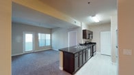 This single-floor space showcases an open-concept design that seamlessly connects the kitchen and living area. The kitchen is a standout feature, equipped with rich, dark wood cabinetry that extends from a peninsula island to upper wall-mounted cabinets. Dark granite countertops provide a striking contrast against the cabinetry, while a double-basin sink sits centrally along the counter run. Stainless steel appliances, including a built-in microwave and range, add a modern, functional touch. The kitchen flooring is finished in light-toned tile, which transitions smoothly into the carpeted living area. The living space is generously proportioned, benefiting from natural light that streams through large windows fitted with vertical blinds. A glass door on the far wall suggests access to an outdoor area, further enhancing the sense of openness. Neutral-toned walls and consistent recessed lighting throughout the space create a clean, cohesive aesthetic. The layout is practical and efficient, making the most of the available footprint while maintaining a comfortable, airy atmosphere throughout.