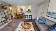 This single-floor residence offers a thoughtfully designed open-concept layout that seamlessly connects the living, dining, and kitchen areas. The living room features dark hardwood-style flooring, a comfortable gray sofa accented with blue throw pillows, and a large wall-mounted television flanked by open shelving units. A glass door fills the space with natural light and provides access to the outdoors. The adjacent dining area is anchored by a round wooden table and cushioned chairs, complemented by a decorative sideboard with a round mirror and matching lamps, creating a warm, inviting atmosphere. The galley-style kitchen is well-equipped with stainless steel appliances, white shaker cabinetry, granite countertops, a tiled backsplash, and a breakfast bar with bar stools. The primary bedroom offers plush carpeting, a large dresser with mirror, and a window with an outdoor view, while the second bedroom features a spacious walk-in closet. The bathroom is finished with tile flooring, a combination tub and shower, and granite countertops. Neutral tones, consistent finishes, and coastal-inspired décor unify the space throughout.