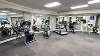 Located at 3901 Indian School Rd NE in Albuquerque, New Mexico, this fitness center offers a dedicated exercise space designed to support a well-rounded workout experience. The facility spans a single floor and is composed of a spacious exercise room and a connecting hallway.

The exercise room is generously sized, featuring a neutral color palette of whites and grays that creates a clean, focused atmosphere. The flooring consists of durable gray carpet tiles throughout, providing comfort and stability during workouts. A drop ceiling with recessed panel lighting ensures the space is evenly and brightly lit.

The room is equipped with a comprehensive selection of cardio and strength-training equipment, including treadmills, elliptical machines, stationary bikes, a rowing machine, and Cybex-branded cable and resistance machines, as well as a weight bench. Large mirrors line multiple walls, visually expanding the space and allowing users to monitor their form. Wall-mounted flat-screen televisions are positioned throughout the room for entertainment during workouts.

A structural column near the center of the room adds an architectural element while maintaining an open, accessible floor plan. The overall layout allows for comfortable movement between equipment, making the space practical and user-friendly for a variety of fitness routines.