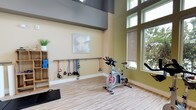 This single-floor fitness studio is thoughtfully designed to support a variety of workout routines in a bright, welcoming environment. The space boasts impressively high ceilings with a two-tone wall treatment — warm beige on the lower half and a cool grey upper section — lending a sense of openness and volume. Large windows along one wall allow abundant natural light to pour in, offering views of surrounding greenery and creating an energizing atmosphere for exercise.

The studio is equipped with spin bikes positioned for easy use, along with a wall-mounted bar rack holding a full assortment of color-coded resistance bands and cable handles, catering to both cardio and strength training needs. A wooden shelving unit provides organized storage for yoga blocks and other accessories, while a step platform on the floor adds versatility for aerobic and functional training exercises. Decorative wall art adds a finishing touch, balancing the functional nature of the space with a clean, modern aesthetic.

The light wood-toned flooring is both durable and visually appealing, complementing the overall design. Roller shades on the windows offer adjustable light control. This fitness studio strikes an ideal balance between practicality and design, making it a motivating space for a wide range of physical activities.