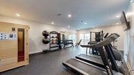 Located at 18217 Cary St in Omaha, Nebraska, this fitness center offers a thoughtfully designed exercise space ideal for a comprehensive workout routine. The room features durable rubber flooring throughout, providing both safety and functionality for a wide range of exercises.

The cardio section is equipped with multiple treadmills and an elliptical trainer, complemented by a wall-mounted television for entertainment during workouts. A full dumbbell rack stocked with a complete set of free weights lines one wall, accompanied by an adjustable weight bench for strength training versatility. A Hoist functional trainer cable machine anchors the strength training area, supporting a broad spectrum of resistance exercises.

Large floor-to-ceiling mirrors span one wall, enhancing the sense of space and allowing for proper form monitoring. Recessed lighting illuminates the room evenly, while a sliding glass door opens directly onto an outdoor deck, introducing natural light and fresh air into the space. A water fountain station is conveniently located near the exit, and the clean, neutral white walls contribute to a bright, motivating atmosphere. The fitness center is well-suited for residents seeking a convenient and fully equipped place to maintain an active lifestyle.