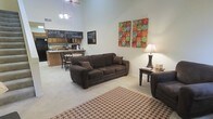 This two-story space makes an immediate impression with its dramatically high ceilings in the main living area, creating a sense of openness and volume that defines the overall ambiance. The open-concept layout seamlessly connects the living room to the kitchen and dining area, promoting a fluid, social atmosphere. The living area is anchored by plush, dark brown upholstered seating arranged around a patterned area rug, complemented by warm lamp lighting and colorful wall art that adds a welcoming, casual character. A side table provides functional surface space alongside the seating arrangement. The kitchen area features wood-toned cabinetry and a breakfast bar with counter seating, making it well-suited for both meal preparation and casual dining. A dining table with chairs sits just adjacent, creating a defined eating space within the open floor plan. Chalkboard signage adds a personalized, informal touch to the kitchen wall. Light-toned carpeting runs throughout the main level, contributing to the warm and relaxed feel of the space. With two floors, the layout offers additional functional space beyond the main living level.