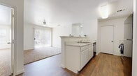 This single-story residence is designed with a clean, bright aesthetic throughout, featuring white walls and a neutral color palette that enhances the sense of openness. The kitchen is positioned centrally and includes a peninsula with a light granite-style countertop, white cabinetry, a built-in sink with a dishwasher, and stainless or black appliances including a refrigerator and range. The kitchen opens directly to the living area, which features wood-look flooring, a ceiling fan, and a sliding glass door with vertical blinds that allows natural light and outdoor access.

The two bedrooms are carpeted and well-proportioned, each featuring windows with horizontal blinds that bring in natural light. One bedroom includes a ceiling fan, adding comfort and ventilation. Both rooms offer clean, versatile spaces suitable for varied furniture arrangements.

Two bathrooms are present in the unit, each featuring wood-look flooring, a shower-tub combination with a curtain, a toilet, and a vanity with white cabinetry. A hallway connects the bedrooms and bathrooms, contributing to a functional and organized layout. The textured ceilings and consistent finishes throughout lend the space a cohesive, well-maintained appearance.