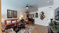 This single-story residence showcases an open-concept floor plan that seamlessly connects the living, dining, and kitchen areas into one cohesive, flowing space. The living area is anchored by rich hardwood-style flooring that extends throughout, complemented by a neutral color palette on the walls and ceilings. A ceiling fan with integrated lighting serves as a functional centerpiece overhead, while recessed lighting provides additional illumination throughout the space.

The living area offers generous room for seating arrangements, as evidenced by the plush sofa and accent chair that frame a dark wood coffee table set atop a vibrant, patterned area rug. Decorative wall art and sculptural accents add personality and visual interest to the space.

Moving deeper into the unit, the layout transitions naturally into a dining area with a table and chairs, positioned adjacent to a kitchen bar counter with barstool seating. The kitchen area, partially visible beyond the pass-through counter, features track lighting and additional cabinetry. The vaulted ceiling detail near the dining area adds architectural dimension, enhancing the overall sense of openness throughout this well-designed residence.