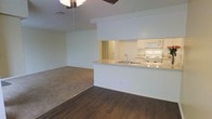 This single-floor residence offers a straightforward, functional layout centered around an open-concept living and dining area. The space features a combination of dark hardwood flooring in the main entry and kitchen zone, transitioning to plush carpeting in the living area, creating a clear visual distinction between the spaces.

The kitchen is designed with practicality in mind, featuring a pass-through counter that opens directly to the living and dining area, promoting a connected, social atmosphere. The kitchen is equipped with a white range, an over-the-range microwave, a double sink, and ample upper cabinetry finished in a light wood tone. The light-colored countertop extends along the pass-through, making it ideal for casual dining or additional prep space.

The living area is generously proportioned and benefits from a ceiling fan that aids in air circulation throughout the space. The neutral white walls provide a clean, versatile backdrop suited to a variety of interior styles. A doorway visible beyond the kitchen hints at additional rooms within the residence. The overall ambiance is light, airy, and practical, offering a welcoming and adaptable environment well-suited for everyday living.