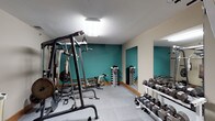 This fitness center is a fully equipped exercise facility designed to support a wide variety of workout routines. The space is divided into distinct functional zones, seamlessly connected within a single floor layout. The strength training area features a multi-station cable machine, a power rack, a dumbbell rack stocked with a full range of free weights, an adjustable bench, and a pull-up/dip station. Large wall-mounted mirrors run along one side, enhancing the sense of space and allowing users to monitor their form. The flooring throughout the strength area consists of interlocking foam tiles, providing cushioning and traction during workouts. The cardio zone houses multiple Precor treadmills, elliptical cross-trainers, and a stationary bike, offering a well-rounded selection of aerobic equipment. The walls are painted in a combination of warm beige and a bold teal accent, giving the space a vibrant yet functional aesthetic. Overhead and wall-mounted lighting provides consistent illumination throughout. An adjacent area with wood cabinetry and a countertop appears to serve as a convenience station. An emergency exit door is also present, ensuring safety compliance. Overall, the facility is thoughtfully organized to accommodate both cardio and strength training needs.