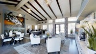 This single-floor space makes an immediate impression with its dramatic vaulted ceilings adorned with bold, dark-stained exposed wooden beams set against crisp white paneling. The soaring ceiling height creates an airy, open atmosphere that is further enhanced by large floor-to-ceiling windows and French doors, which flood the interior with natural light and offer views of the surrounding outdoor greenery. Geometric pendant chandeliers in a matte black metal finish hang from the ceiling, adding a contemporary yet sophisticated touch to the space. The main lounge area is thoughtfully arranged with white upholstered armchairs featuring nailhead trim detailing, centered around a dark circular cocktail table atop layered area rugs in soft, neutral tones. A reception or service counter area is visible toward the rear of the space, finished in dark wood tones to complement the beams above. Patterned drapery in black and white geometric prints frames the windows, adding visual depth and contrast. Curated wall art, decorative mirrors, and sculptural accents contribute to the polished, modern aesthetic throughout. Dark hardwood flooring ties the entire design together, creating a cohesive and inviting environment that balances elegance with comfort.