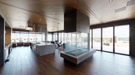 Located at 351 S Jackson St in Denver's vibrant Washington Park neighborhood, this contemporary single-floor amenity space offers a seamlessly integrated open-plan design that blends indoor comfort with outdoor living. The interior is anchored by a striking, glass-enclosed double-sided fireplace with a dark brick chimney column, serving as a dramatic architectural centerpiece that divides the lounge and gathering areas while maintaining visual connectivity throughout.

The gourmet kitchen is a standout feature, equipped with a large concrete-topped island, professional-grade gas cooktop, double wall ovens, stainless steel range hood, and open shelving set against textured blue-gray brick accent walls. Rich, wide-plank hardwood flooring runs throughout the space, complemented by warm wood-paneled ceilings that add depth and a refined natural aesthetic.

Multiple lounge seating areas are thoughtfully arranged to take advantage of floor-to-ceiling glass walls, which frame expansive city views of the Denver skyline and surrounding neighborhood. Sliding glass doors provide direct access to a generous outdoor terrace, outfitted with additional seating, landscaped planters, and string lighting—ideal for gatherings at any time of day. Exposed concrete columns punctuate the interior, reinforcing the space's modern industrial character while adding structural interest.