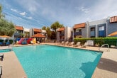 Located at 5150 W Eugie Ave in Glendale, Arizona, this outdoor amenity area is centered around a generously sized swimming pool with crystal-clear turquoise water. The pool area is designed for both relaxation and recreation, offering a separate spa alongside the main pool. A wide, textured concrete pool deck surrounds the water and is furnished with multiple lounge chairs and chaise lounges, ideal for sunbathing in the Arizona climate. A covered gazebo provides a shaded retreat within the space. One of the most distinctive design elements is a series of bold, colorful murals featuring abstract geometric shapes in vibrant reds, oranges, blues, and yellows, adding an artistic and contemporary character to the environment. The complex's residential buildings frame the pool area, showcasing a Southwestern architectural style with stucco exteriors, terra-cotta tile rooflines, and balconies. Mature trees and manicured green hedges line the perimeter, offering natural shade and a lush contrast to the desert surroundings. The pool area is enclosed by a wrought iron fence, ensuring privacy and security. The overall atmosphere is bright, resort-inspired, and well-suited to the warm Glendale, AZ climate.