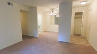 This single-floor space features an open-concept layout that connects the living area to the kitchen, creating a sense of continuity throughout. The living area is finished with neutral-toned wall-to-wall carpeting, lending a comfortable and understated feel to the space. Structural columns define the boundaries between zones without fully enclosing them, maintaining an airy, open quality.

A ceiling fan with a light fixture is centrally positioned overhead, providing both ambient lighting and air circulation. The walls are painted in a soft, light neutral tone that contributes to the bright and clean atmosphere of the interior.

The kitchen is partially visible through the opening and features white appliances, including a range and oven, along with white cabinetry. A pass-through counter connects the kitchen to the adjacent dining or living area, allowing for easy interaction between spaces. The kitchen flooring transitions from the carpeted living area to a hard surface, adding a practical distinction between the two zones.

To the right, a doorway hints at additional rooms beyond, suggesting the layout extends further. Overall, the space presents a functional and straightforward design with a clean, move-in ready aesthetic.