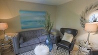 This two-story space offers a well-appointed interior with a comfortable and stylish living area. The sitting room showcases neutral-toned walls that provide a calm backdrop, complemented by a plush, tufted gray sofa that anchors the seating arrangement. A sleek, dark leather wingback chair adds a sophisticated contrast, dressed with a textured faux-fur accent pillow for a cozy touch. A large, abstract canvas in soft aqua and teal tones serves as a striking focal point on the wall, introducing a serene, coastal-inspired palette to the room. A bold blue glass vase filled with tall, feathery greenery adds organic texture and visual height to the space, while a sculptural white decorative piece on the floor contributes an artistic element. The room is grounded by a geometric patterned area rug in neutral tones, adding depth and dimension underfoot. The overall ambiance is relaxed yet polished, blending contemporary design elements with warm, inviting accents. The two-floor layout suggests additional living space beyond what is visible, offering ample room for varied functions throughout the home.
