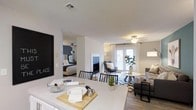 This single-floor residence showcases an open-concept design that seamlessly integrates the kitchen, dining, and living spaces into one cohesive and functional area. The kitchen features a generous light-colored countertop that serves as both a prep space and a casual gathering point, flowing naturally into the dining area, which is furnished with a sleek table and black chairs. A fireplace serves as a focal point in the central living zone, adding warmth and architectural interest to the space. The living area is thoughtfully arranged with a sectional sofa, patterned area rug, and accent chairs, creating a welcoming environment. French doors at the far end of the room invite natural light throughout and offer access to the outdoors. Ceiling fans are present in multiple zones, supporting air circulation and comfort. Wood-look flooring runs through the main living spaces, contributing a warm and contemporary aesthetic. A peek into the adjacent bedroom reveals a well-styled sleeping area with its own ceiling fan, completing the home's cohesive and modern interior design.