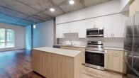 Unit 135 at 219 9th Ave is a 680-square-foot, one-bedroom, one-bathroom apartment in Oakland, CA, showcasing a thoughtfully designed industrial-modern aesthetic throughout its single floor. The open living area features rich hardwood-style flooring, exposed concrete ceilings, and large windows that invite natural light and offer views of the surrounding greenery. The well-appointed kitchen is a standout feature, boasting two-tone cabinetry with white upper cabinets and warm wood-tone lower cabinets, a white subway tile backsplash, stainless steel appliances including a refrigerator, range, microwave, and dishwasher, and a movable kitchen island. The 140-square-foot bedroom is carpeted for comfort, features an exposed concrete ceiling, and includes a large window with outdoor views and access to a dedicated walk-in closet with built-in hanging rods and plush carpet. The bathroom is finished with large-format light tile flooring, a full soaking tub and shower combination, and clean white cabinetry with ample counter space. Concrete structural columns and ceilings are preserved throughout, lending the unit an urban, loft-inspired character that is balanced by warm wood tones and crisp white walls.